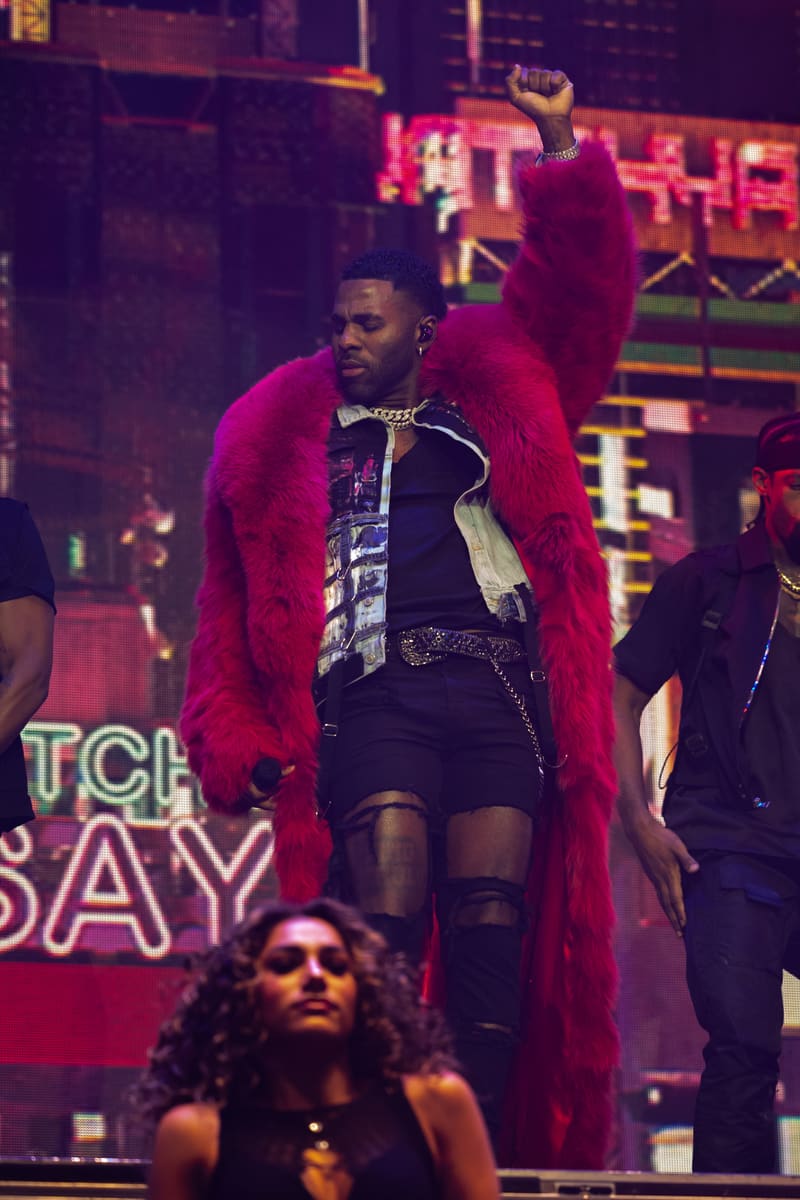 Performer in a striking magenta coat and ripped black trousers on stage, bathed in neon purple lighting.