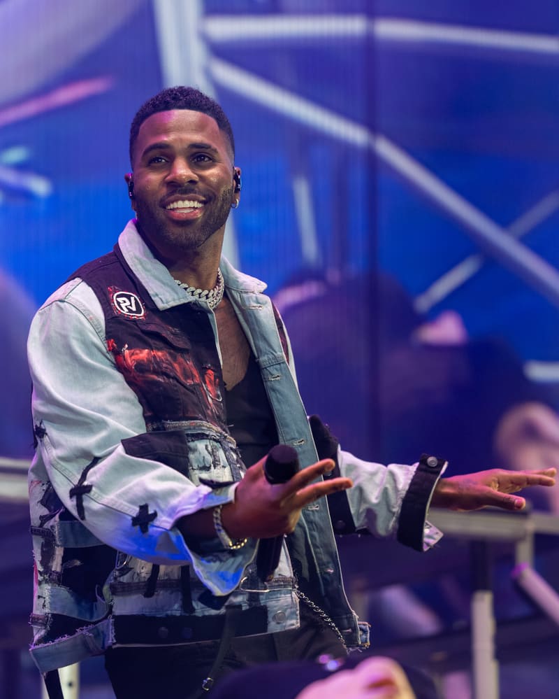 Performer in a patterned shirt and vest gestures expressively on stage under blue lighting.