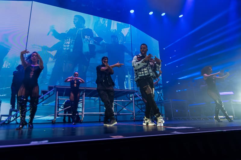 Performer in checked jacket centre stage with band members and large video screen backdrop under blue stage lighting.