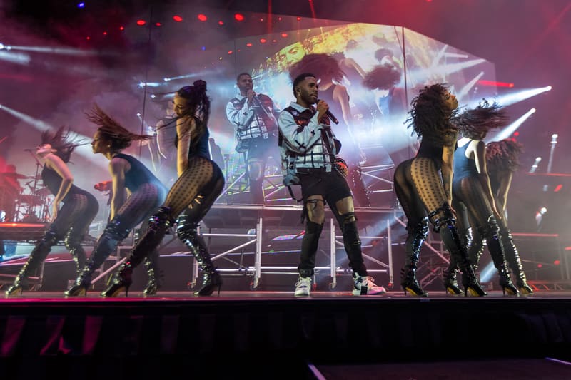 Performer centre stage surrounded by dancers in coordinated outfits under dynamic purple and pink stage lighting.