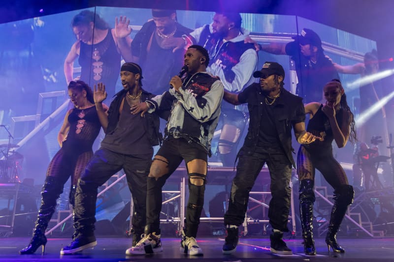Performer in white jacket and black pants dancing centre stage with backing dancers under purple stage lighting.
