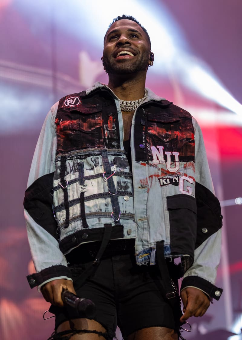 Performer on stage wearing a patchwork denim jacket and chains, smiling beneath purple and pink stage lighting.