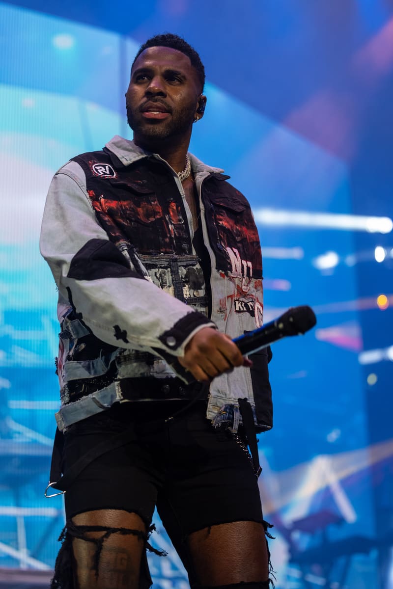Performer on stage holding a microphone, wearing a patterned black and grey jacket and ripped shorts, against a blue-lit backdrop.