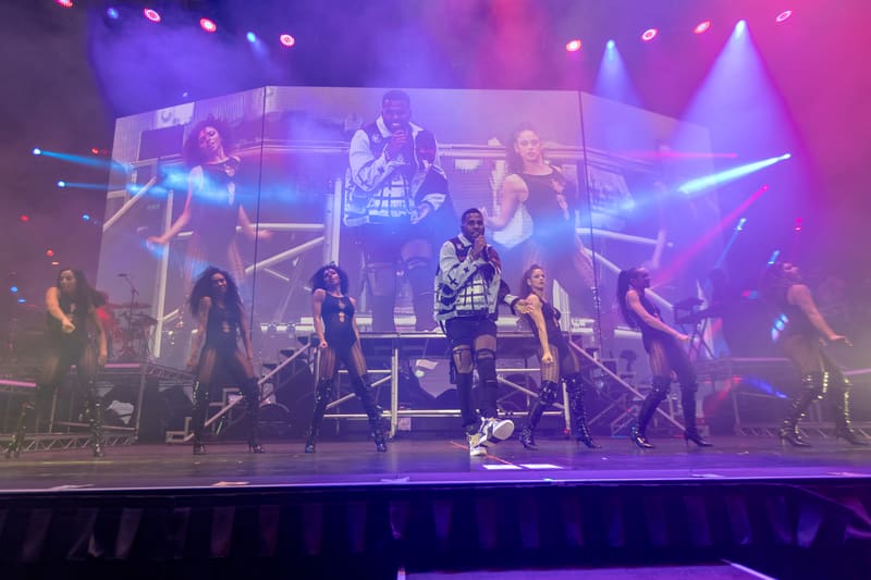 Performers on an elevated stage lit by purple and blue spotlights during a concert production.