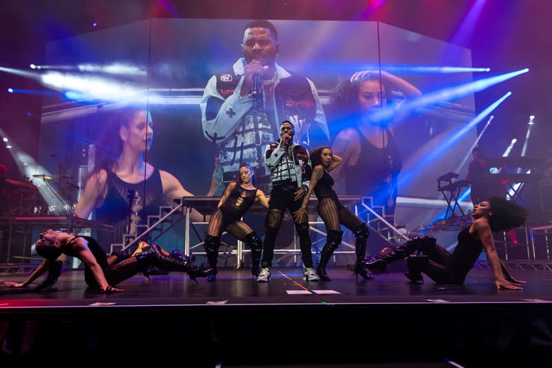 Dancers perform on stage with a large screen projection of a performer above them, lit by blue and purple stage lights.