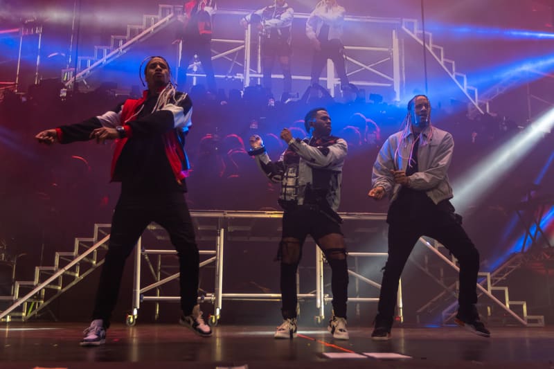 Four dancers perform on stage under blue and purple spotlights with geometric set design behind them.