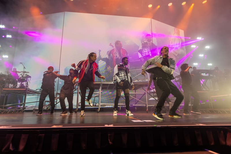 Performer in red jacket dancing centre stage with backing band under purple and pink stage lighting.