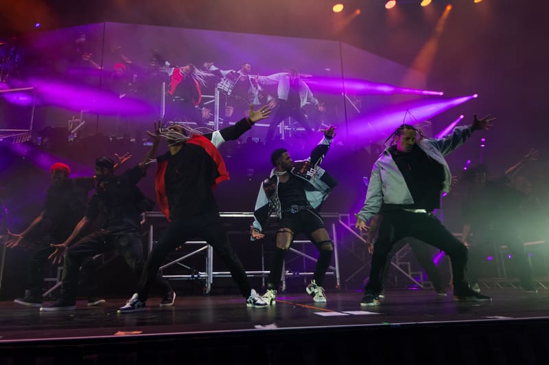 Live performers dance and move energetically on stage under vibrant purple stage lighting during a concert performance.