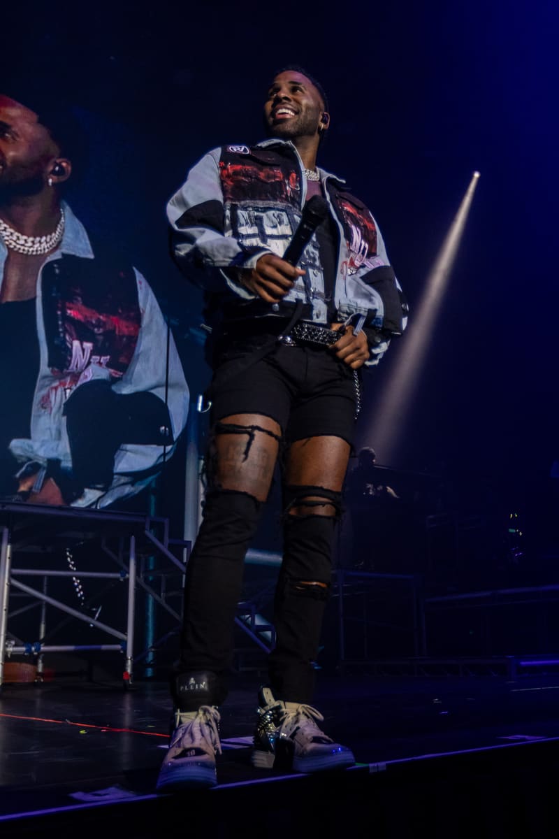Performer on stage in graphic jacket and ripped black shorts, lit by blue spotlights, gesturing to audience.