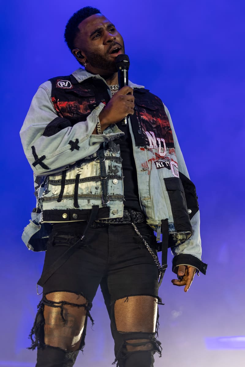 Performer on stage holding a microphone, wearing a patterned jacket and ripped black shorts against blue stage lighting.