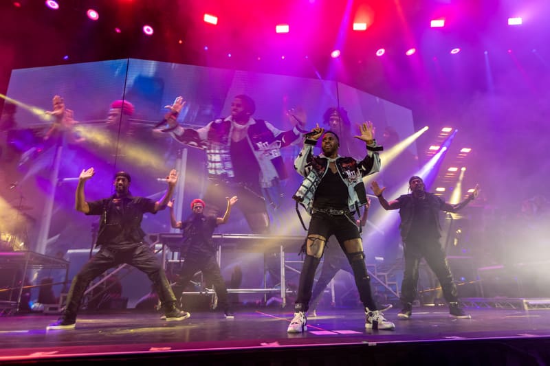 Performer in studded black jacket centre stage with dancers and band members under vibrant purple and pink stage lighting.