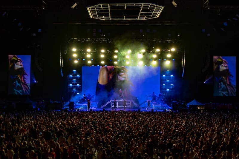 Performer on stage beneath bright lights with large crowd and illuminated screens at concert venue.
