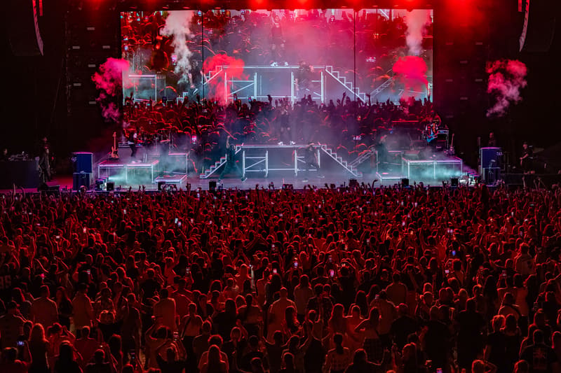 Massive crowd with raised hands beneath illuminated stage screens glowing red and blue at a concert venue.