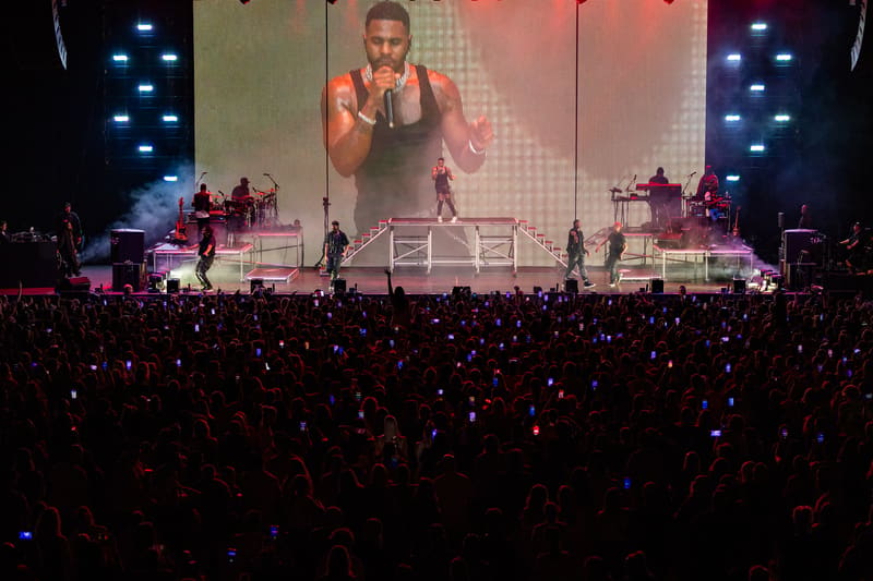 Performer on stage with large video screen behind, surrounded by blue stage lighting and crowd holding up phones.