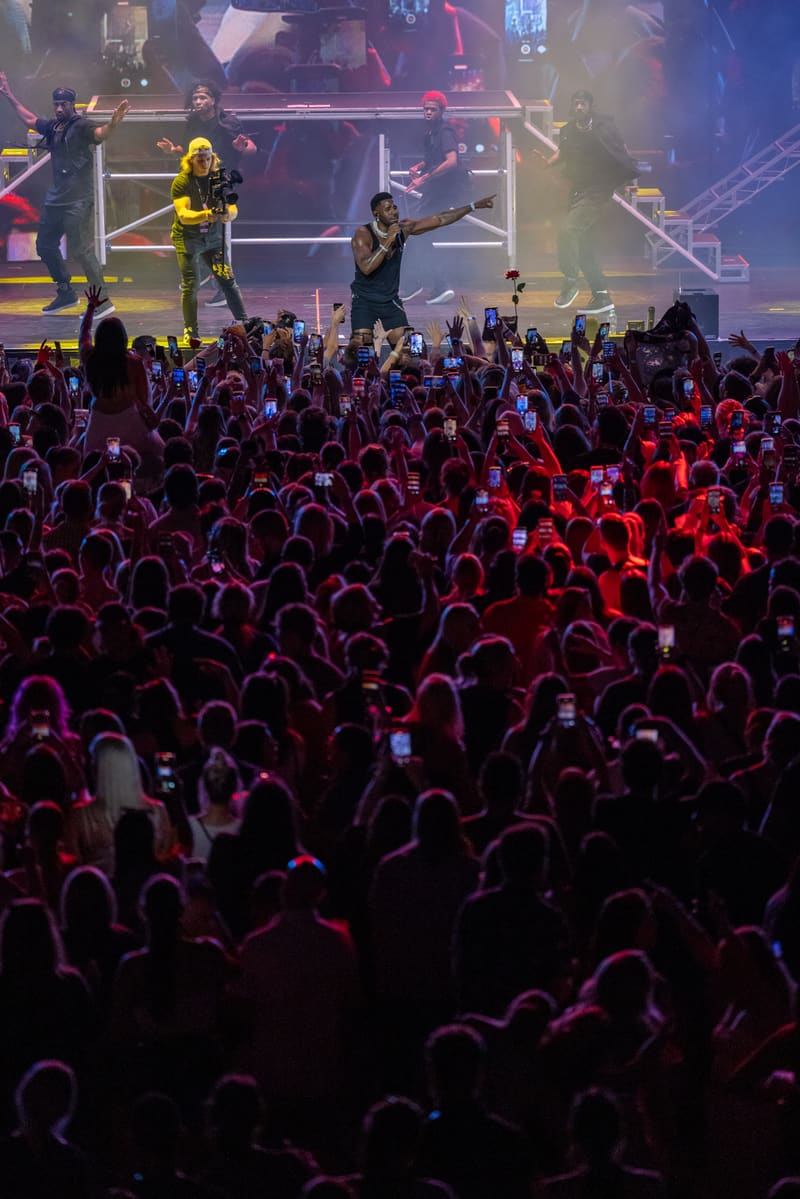 Performers on an elevated stage lit by red and purple spotlights engage an energetic crowd at a concert venue.