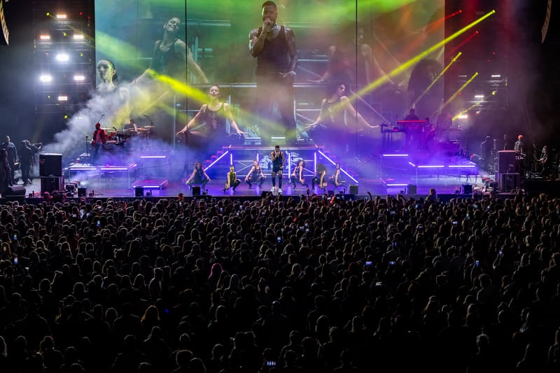 Performer on stage with vibrant purple and green lighting above a large crowd at a concert venue.