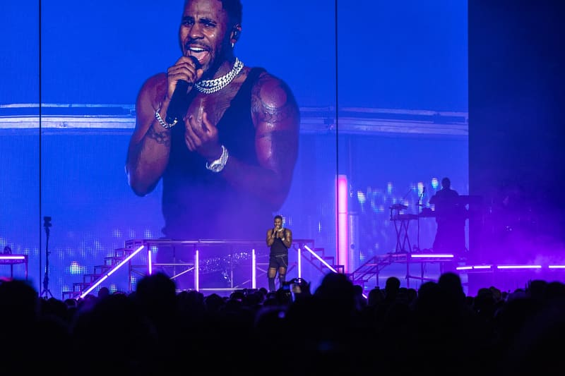 Performer on stage with large video screen backdrop and purple lighting, silhouetted crowd in foreground.