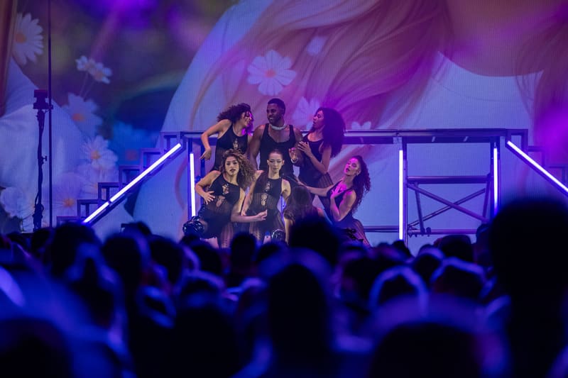 Performers on stage beneath purple and blue lighting with audience silhouettes in foreground.