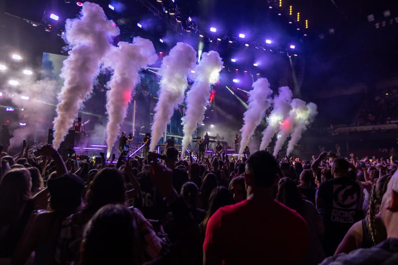 Crowd of concertgoers with hands raised as purple and white pyrotechnics explode across the stage.
