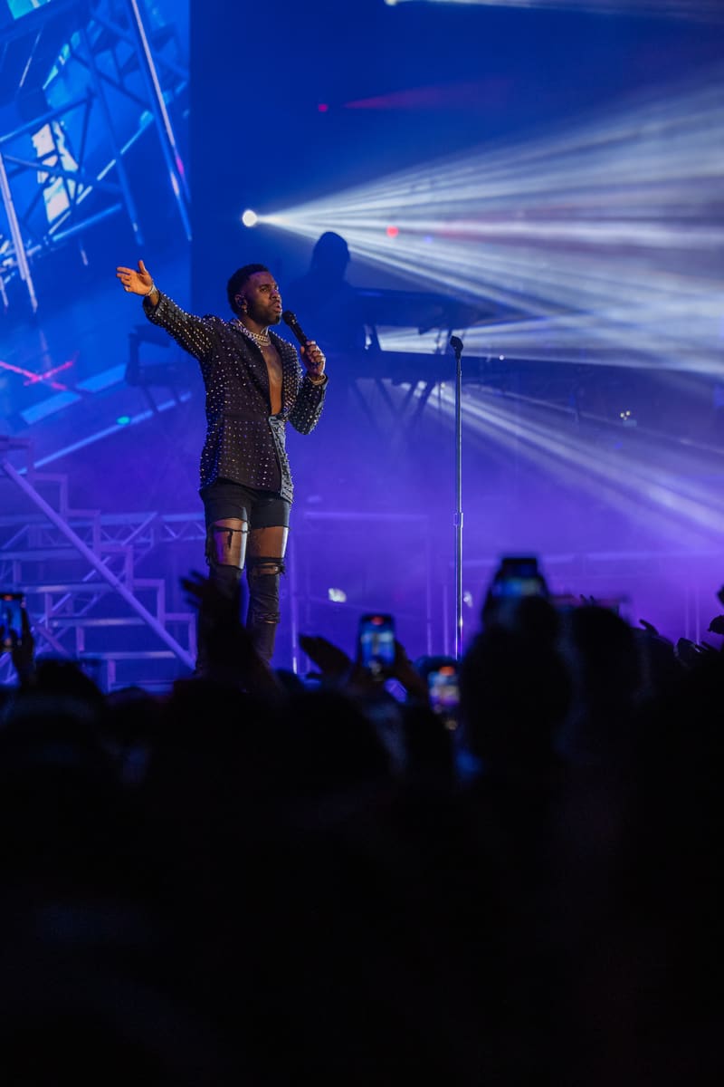 Performer on stage in a patterned jacket with arm raised, bathed in blue and purple stage lighting while audience members record on phones below.