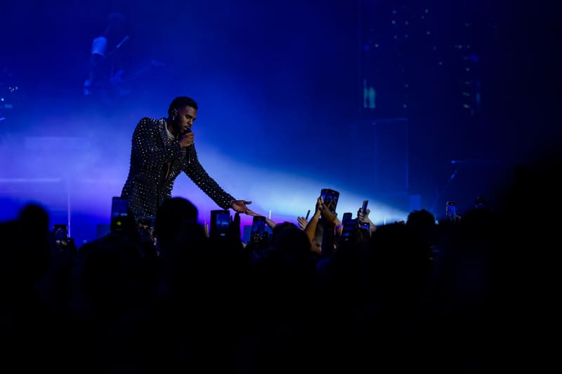Performer on stage bathed in blue light, reaching towards audience silhouettes in foreground.