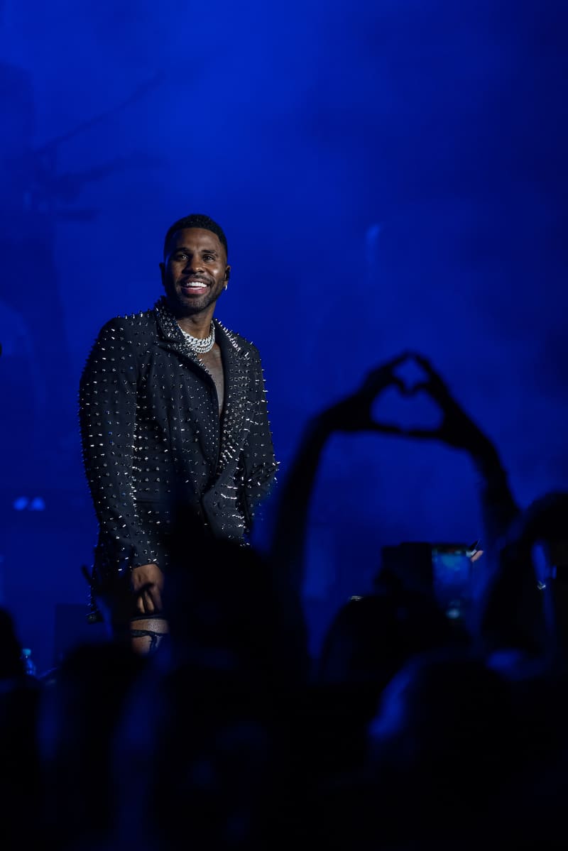 Performer on stage wearing a studded black jacket, illuminated by blue stage lighting while audience members hold up phones.