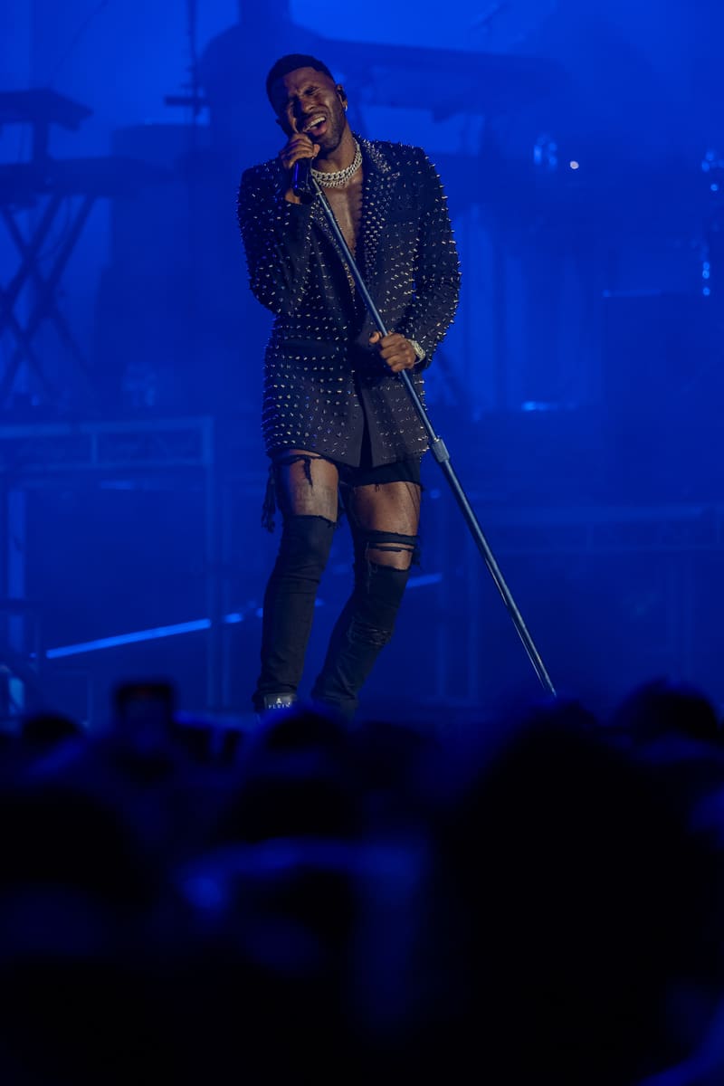 Performer on stage in studded jacket and ripped shorts, singing into microphone under blue stage lighting.