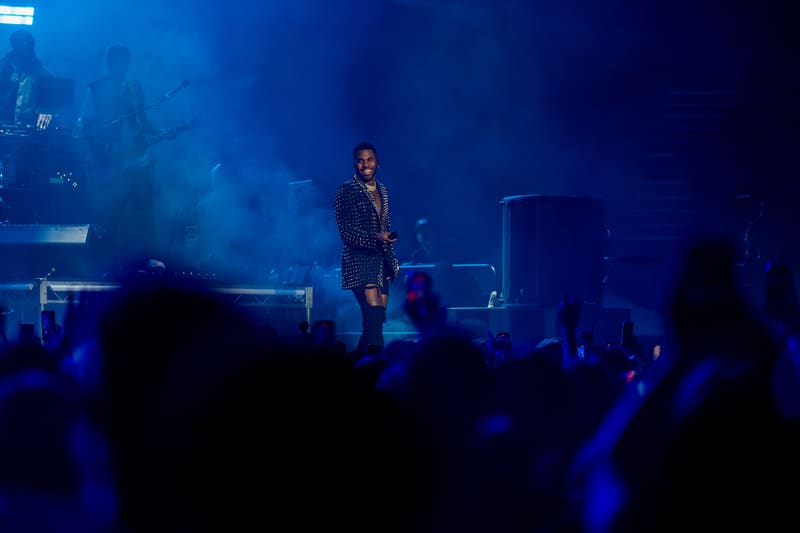Performer on stage bathed in blue lighting, silhouetted crowd in foreground with raised hands.