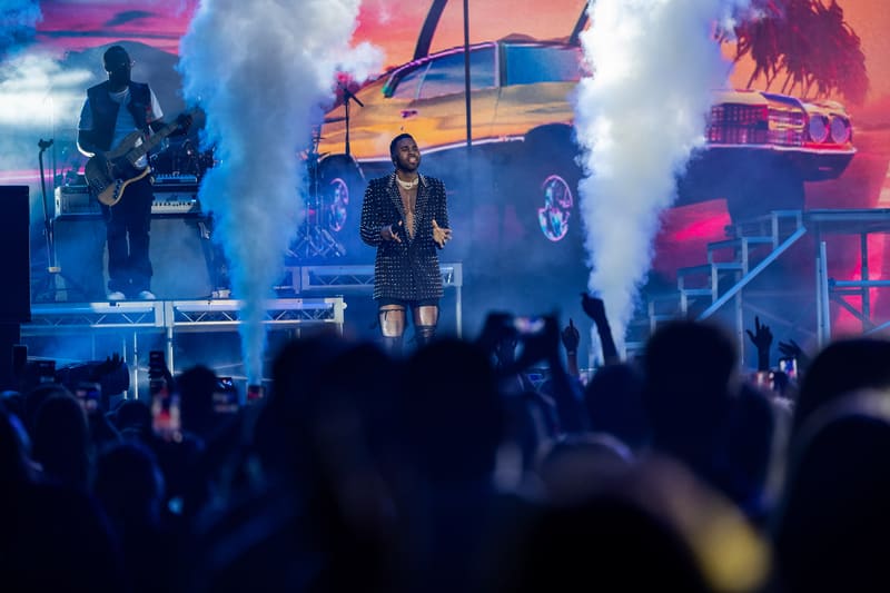 Performer on stage surrounded by pyrotechnic flames and smoke with animated vehicle graphics projected behind.