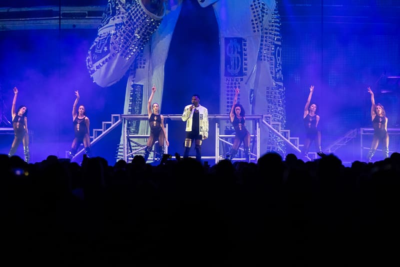 Performer centre stage under blue spotlights with backing dancers and large video screens during a concert production.
