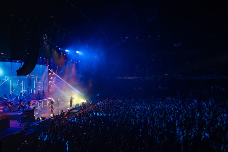 Packed concert venue with stage illuminated by blue spotlights and large crowd in darkness.