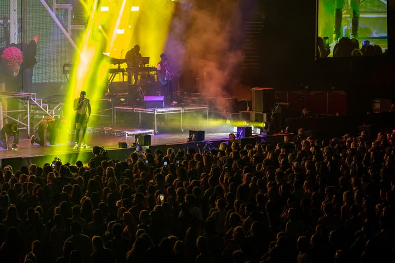 Performer on stage under bright yellow and purple lighting with a large crowd in the foreground at a concert venue.