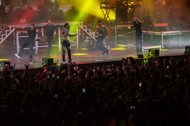 Performer on stage with dancers under bright yellow and red lighting, surrounded by a crowd of audience members holding up phones.