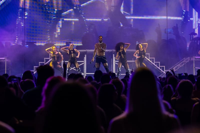 Performer and backing dancers on an elevated stage beneath purple lighting, surrounded by an enthusiastic crowd.