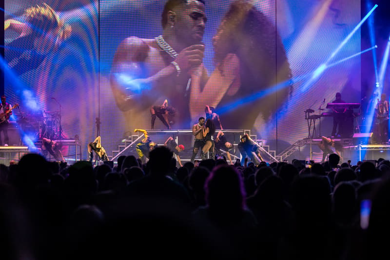 Performer on stage backed by large video screens and blue stage lighting, with audience silhouettes in foreground.