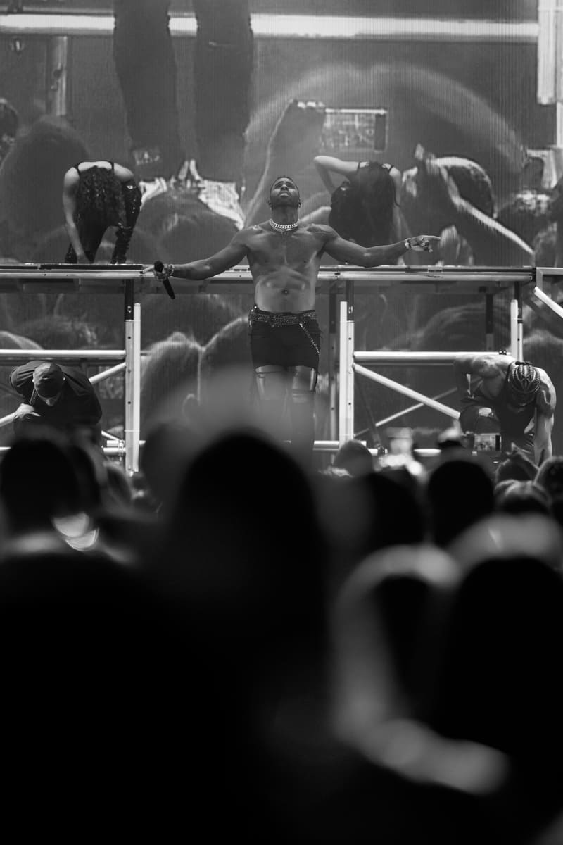 Shirtless performer with arms outstretched on stage, surrounded by backing dancers and cheering crowd in black and white.