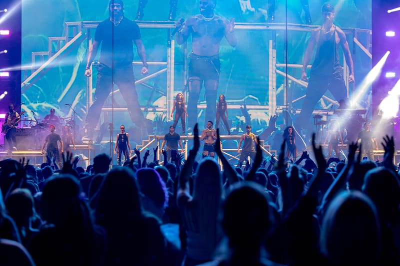 Crowd with raised hands silhouetted against a brightly lit stage with blue and purple lighting effects.