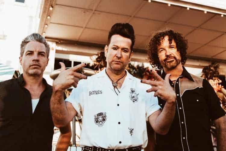 Three band members pose together in casual attire, arms raised in a relaxed group gesture.