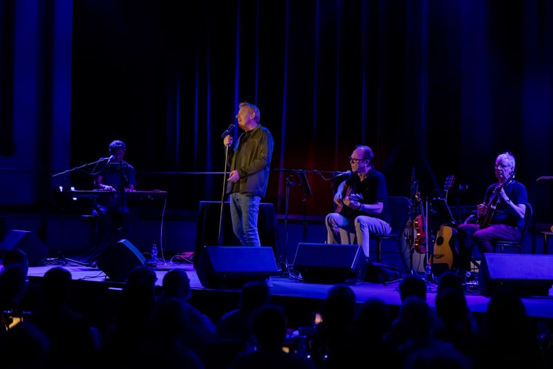 Four musicians perform on a dimly lit stage with blue atmospheric lighting, featuring vocals and acoustic instruments.