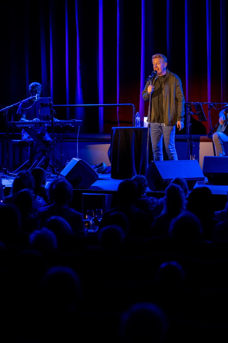 Singer performing centre stage with microphone, accompanied by pianist, beneath vertical blue neon lighting, audience silhouetted in foreground.