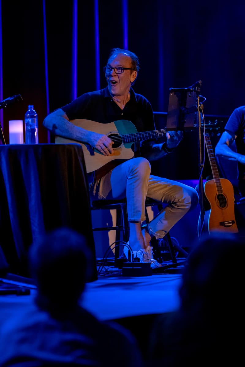 Guitarist seated on stage with acoustic guitar, bathed in blue stage lighting, performing before an audience.