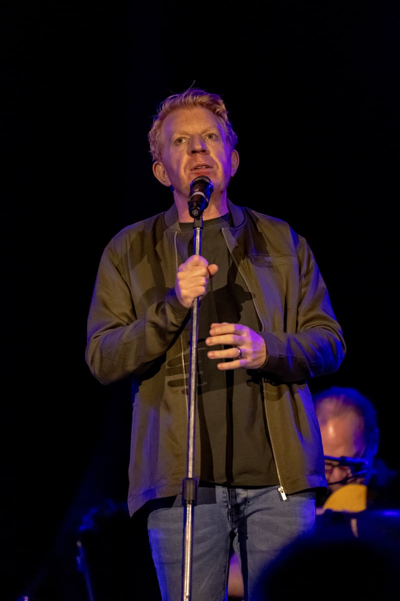 Singer in olive jacket performing at microphone under purple stage lighting.