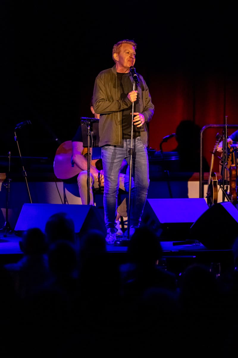Singer in brown jacket holding microphone on stage lit by blue and purple spotlights, audience silhouetted in foreground.