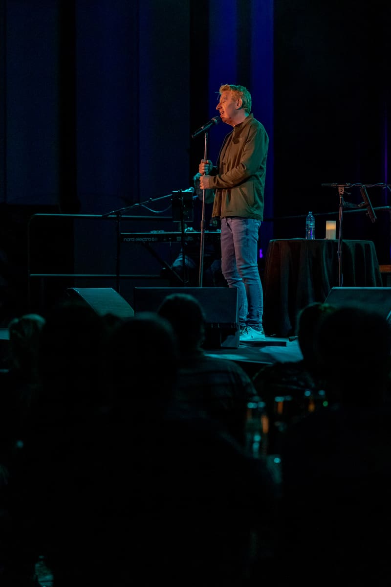 Male performer stands at microphone on dimly lit stage with keyboard and audience silhouettes in foreground.