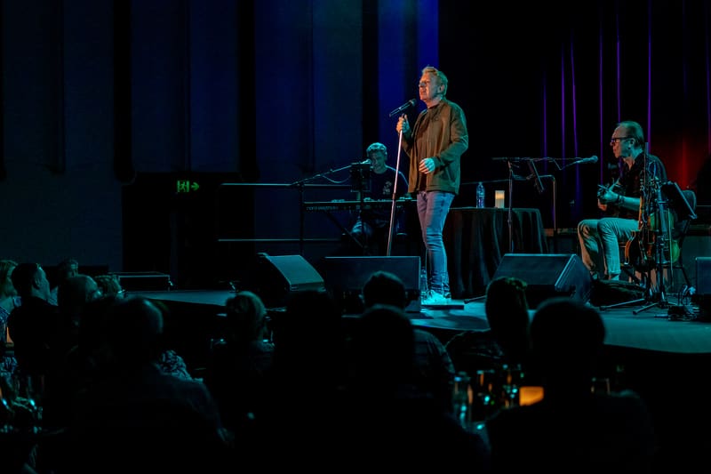 Male performer in turquoise-lit stage setting with pianist, audience silhouettes in foreground.