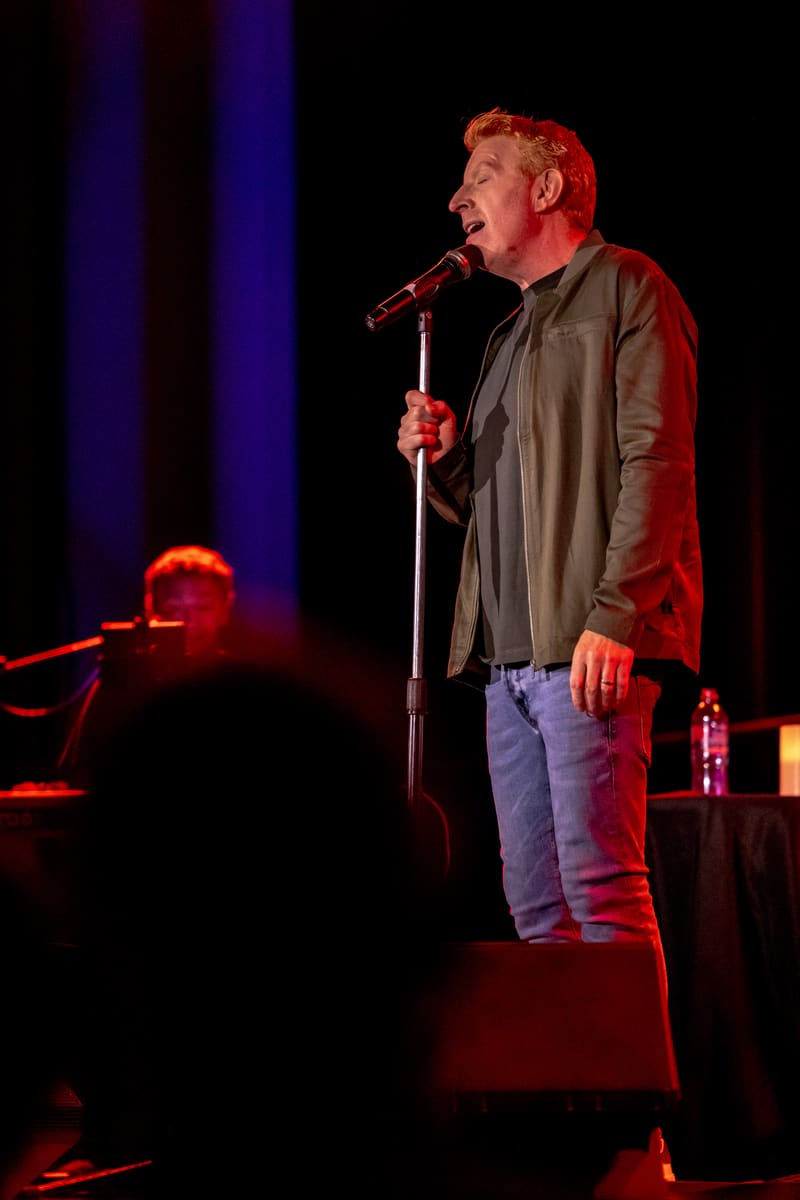 Male performer singing at microphone on stage under warm red and blue stage lighting.