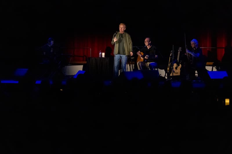 Male performer centre stage with band members and instruments bathed in warm stage lighting against a dark background.
