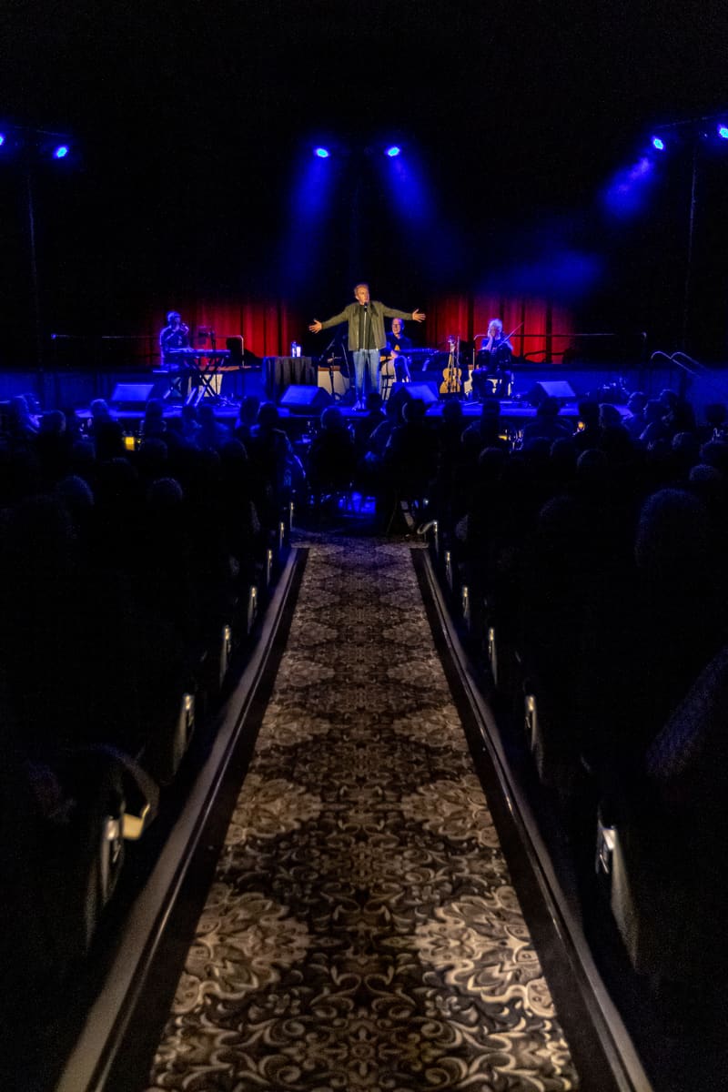 Singer in dark suit stands centre stage under blue spotlights, addressing seated audience along a patterned runway.