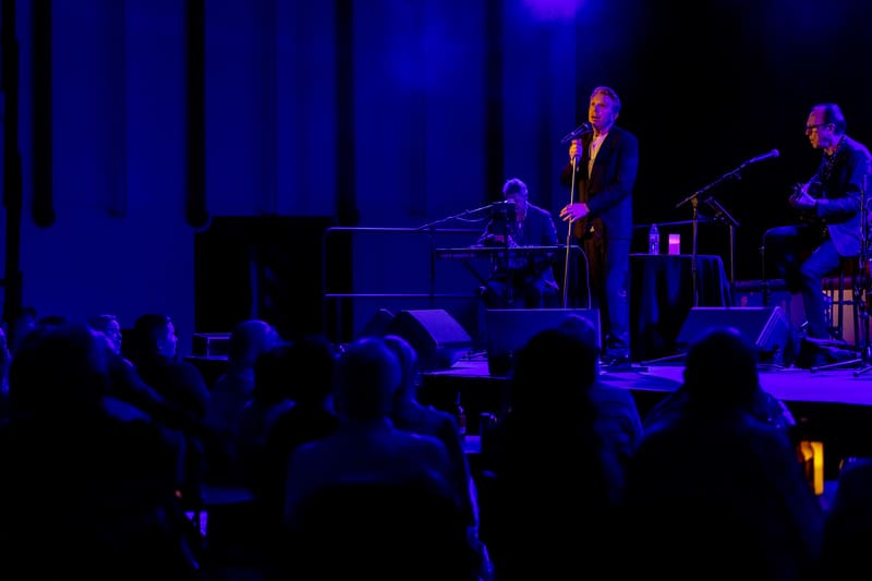 Performer in dark suit standing at microphone on stage with pianist and band members, blue stage lighting illuminating the scene.