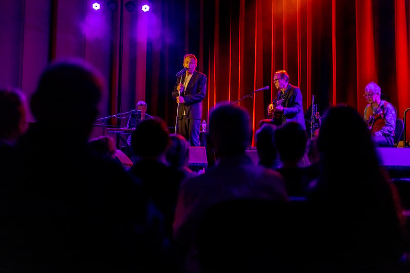 Musicians perform on stage bathed in purple and red lighting, with audience silhouettes in the foreground.
